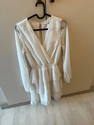 Vit studentklänning från Bubbleroom - Säljer en vit omlottklänning från Bubbleroom med långa ärmar och volangdetaljer. Klänningen har v-ringning, markerad midja och flera lager volanger i kjolen för en flowy look. Amvänt en gång.