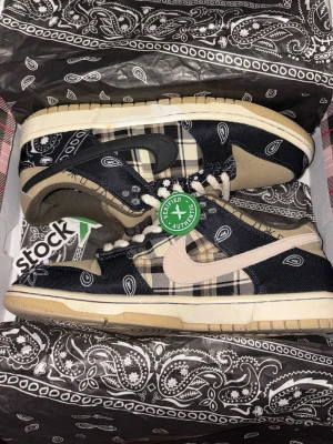 Nike SB Dunk Low Travis Scott Paisley - Unika Nike SB Dunk Low Travis Scott sneakers med paisley och rutigt mönster i svart, beige och grått. Skorna har tjocka vita snören, beige sula och detaljer i mocka. Swoosh-logga på sidan och 'Cactus Jack' branding på plösen.
