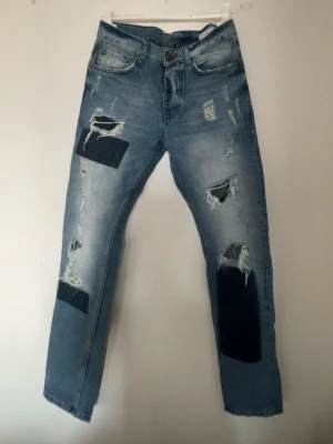 Blå patchade jeans Adrian Hammond - Säljer ett par blå jeans från Adrian Hammond med slitningar och svarta patchar på benen. sopiga detaljer och en relaxed vibe. Perfekta för dig som gillar en edgy streetstil. Modellen är Boyle och materialet är klassisk denim.