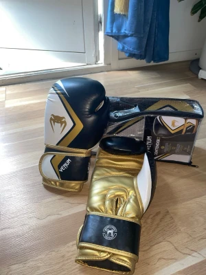 Venum svarta och guld boxningshandskar - Snygga boxningshandskar från Venum i svart, vitt och guld. Handskarna har ett coolt ormlogotyp på ovansidan och bred kardborrestängning för extra stöd. Tillverkade i slitstarkt syntetmaterial, perfekta för träning och sparring.