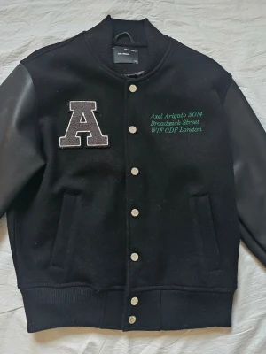 Svart varsity overshirt Axel Arigato - Snygg svart varsity overshirt från Axel Arigato med stora A-patchen på bröstet och grön brodyrtext. Jackan har tryckknappar, ribbade muddar och kontrasterande ärmar i skinnimitation. Storlek S, men sitter som Medium. 