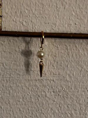 Guldörhänge med pärla och spike - Coolt örhänge i guldfärgad metall med en rund vit pärla och en spetsig spike-hänge. Modern design som sticker ut och passar perfekt för dig som gillar unika accessoarer. Säljs styckvis.