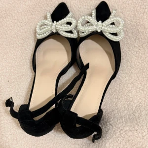 Svarta pumps med pärlrosett och klack - Snygga svarta pumps i mocka-look med spetsig tå och bred klack. Skorna har en dekorativ rosett framtill i pärlor och knytband runt vristen för extra detalj. Perfekta för dig som vill sticka ut med en elegant och trendig stil.