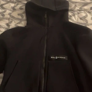 Svart hoodie från Sail Racing M - Svart hoodie från Sail Racing med dragkedja framtill och huva. Klassisk logga på bröstet och stora fickor. Perfekt för dig som gillar stilren och sportig look. Skön passform och enkel att matcha med det mesta.