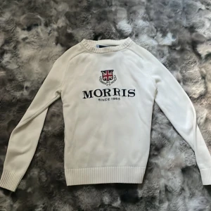 Vit stickad tröja från Morris - Snygg vit stickad tröja från Morris med broderad logga och brittisk flagga på bröstet. Tröjan har långa ärmar, ribbade muddar och en svart siffra 8 på ena ärmen. Perfekt för dig som gillar klassisk och stilren design.