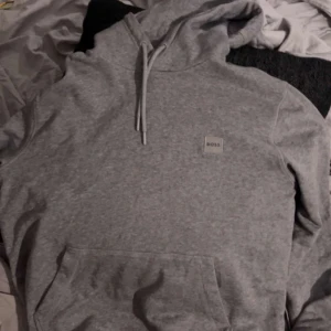 Boss Hoodie - Säljer en stilren grå hoodie från BOSS med klassisk huva och snörning. Tröjan har en liten BOSS-logga på bröstet och en stor magficka framtill. Perfekt för dig som gillar enkel och clean streetstyle.