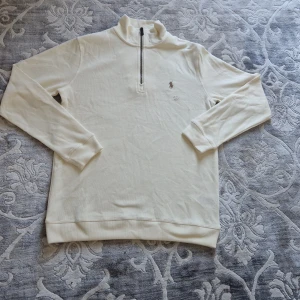 Vit half zip tröja Polo Ralph Lauren - Vit långärmad tröja från Polo Ralph Lauren med klassisk broderad logga på bröstet. Tröjan har en hög krage och half zip-dragkedja framtill. Tillverkad i mjuk bomull och har en clean, stilren look som passar perfekt till jeans eller chinos.