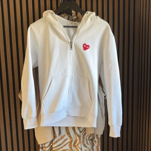 Vit hoodie Comme des Garçons Play  - Vit hoodie från Comme des Garçons Play med dragkedja, huva och känguruficka. Ikoniskt rött hjärta broderat på bröstet. Mjuk bomullskvalitet och klassisk passform, perfekt för en clean och stilren look. Passar nog XS också. Den är lite liten 
