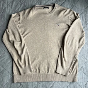 Beige stickad tröja från GANT - Klassisk beige stickad crewneck från GANT med diskret logga broderad på bröstet. Tröjan är i bomull, har ribbade muddar och lång ärm. Väldigt schysst.