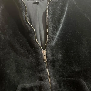 Svart teddyjacka med huva och dragkedja - Mysig svart teddyjacka med huva och stora fickor framtill. Jackan har en rak passform och stängs med dragkedja. Perfekt för kyliga dagar när du vill vara både varm och stilren.