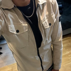Beige vindjacka från Stone Island - Säljer en beige vindjacka från Stone Island med klassisk patch på ärmen. Jackan har två stora bröstfickor med knappar, hög krage och dragkedja framtill. Materialet är lätt och polyester, perfekt för vår och höst. Passformen är normal och jackan är långärmad. Passar S men är storlek junior