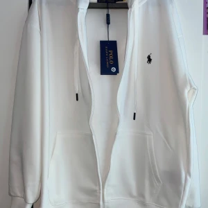 Vit hoodie från Polo Ralph Lauren - Vit hoodie från Polo Ralph Lauren med dragkedja, huva och snörning. Klassisk design med broderad logga på bröstet och två stora fickor framtill. Tillverkad i mjuk bomull för en skön och avslappnad känsla. 