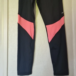 Röhnisch Shape Aine 7/8 Tights - Snygga svarta träningstights från Röhnisch med rosa detaljer över låren. Tightsen har en åtsittande passform och är tillverkade i ett stretchigt syntetmaterial som passar perfekt för träning. Det är lite missfärgat på det rosa på byxorna insida bak.