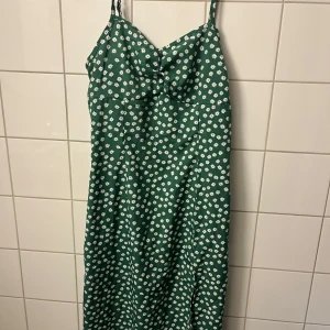 Grön blommig långklänning - Snygg grön långklänning med vita blommor och smala axelband. Klänningen har en feminin v-ringning och figurnära passform upptill som ger en fräsch och somrig vibe. Perfekt för dig som gillar blommigt och vill sticka ut med färg.