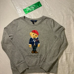 Grå sweatshirt Polo Bear Ralph Lauren - Superfin grå sweatshirt från Polo Ralph Lauren med ikoniska Polo Bear-trycket framtill. Tröjan har rund hals, långa ärmar och mjukt material i bomullsmix. Perfekt för dig som gillar klassisk men lekfull stil. Snygg detalj med björnen i röd basker och blå kavaj. Xs/L barn