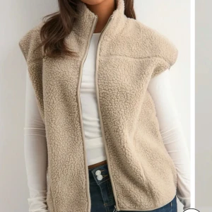 Beige fleeceväst med dragkedja - Mysig beige fleeceväst med hög krage och hel dragkedja framtill. Västen har en avslappnad passform och är tillverkad i mjukt teddyfleece-material som ger en trendig och chill look. Perfekt att slänga över en långärmad tröja för extra värme.