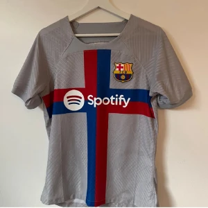 FC Barcelona grå bortatröja Nike - Snygg FC Barcelona bortatröja från Nike i grått med röd och blå korsdesign på framsidan. Tröjan har klubbmärke, Spotify-logga och korta ärmar. Tillverkad i lätt och ventilerande funktionsmaterial, perfekt för fotboll eller att bära som supporter.