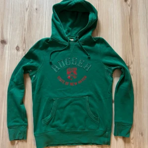 Grön hoodie från Gant - strl XS - Snygg grön hoodie från Gant med trycket 'Rugger State of New Haven' och röd sköld på bröstet. Tröjan har huva med snörning, känguruficka och långa ärmar. Perfekt för dig som gillar klassisk college-stil.