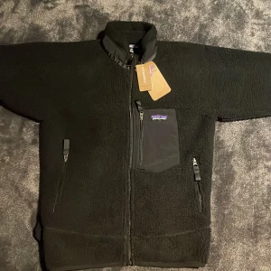 Svart teddyjacka från Patagonia M - Svart teddyjacka från Patagonia med hög krage, dragkedja framtill och tre fickor med dragkedja. Jackan har en bröstficka i kontrasterande material och klassisk Patagonia-logga. Perfekt för kalla dagar och riktigt skön att bära.