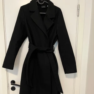 Svart kappa med bälte från Vero Moda  - Stilren svart kappa från vero Moda med klassisk krage och knytskärp i midjan. Lång modell med raka ärmar och snyggt fall. Perfekt för dig som vill ha en tidlös och enkel look.