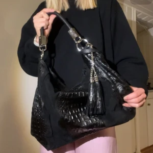 Svart axelväska i skinn med tofs - Snygg svart axelväska från Borse in Pelle, tillverkad i äkta skinn med croco-mönstrade detaljer och dekorativ tofs i metallkedja. Väskan har ett rejält handtag och cool struktur som ger en lyxig vibe. du kommer ha plats för dator i väskan, då den har en brädd på 42 cm.Perfekt för dig som vill ha en statement-väska med italiensk känsla.