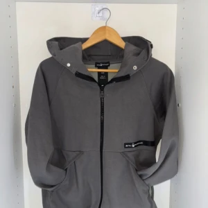 Grå hoodiejacka från Sail Racing - Snygg grå hoodiejacka från Sail Racing med dragkedja framtill och praktisk huva med tryckknappar. Jackan har två stora fickor och en diskret logga på bröstet. Perfekt för chill dagar och enkel att matcha med jeans eller joggers.