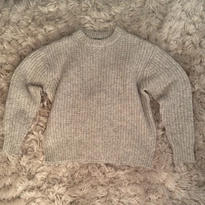 Grå stickad tröja från 157 - Mysig grå stickad tröja från 157 med högre rund halsringning och grovstickad struktur. Perfekt för lager-på-lager och chill dagar. Tröjan har långa ärmar och breda muddar vid ärmslut och nederkant.