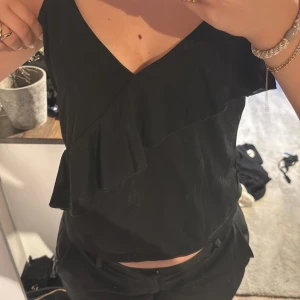 Svart linnetopp med volang från H&M - Svart linnetopp från H&M med v-ringning och breda axelband. Toppen har en snygg volangdetalj framtill som ger en cool och trendig look. Perfekt att styla med både jeans och kostymbyxor. Materialet känns mjukt och lätt.
