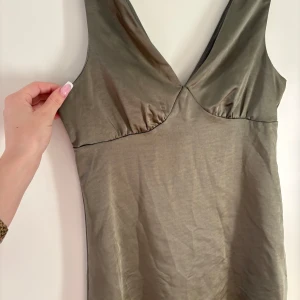 Olivgrön ärmlös klänning med v-ringning - Supersnygg olivgrön klänning med djup v-ringning och ärmlös design. Klänningen har en lätt glansig yta och en enkel, rak passform som ger en stilren look. Perfekt för dig som gillar minimalistiska och trendiga plagg.