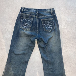 Blå bootcut jeans med broderade fickor - Snygga blå jeans med bootcut passform och broderade fjärilar på bakfickorna. Jeansen har klassisk femficksdesign, midja under naveln och är tillverkade i slitstarkt denim. Perfekta för dig som vill ha en cool och unik detalj på dina jeans.
