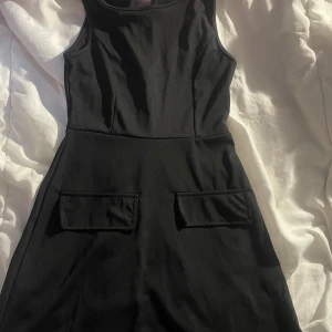 Svart ärmlös kortklänning XS - Svart ärmlös kortklänning i stretchigt material med två fickor framtill. Klänningen har en figurnära passform upptill och lätt utställd kjol. Tillverkad i 95% polyester och 5% spandex för en bekväm känsla.