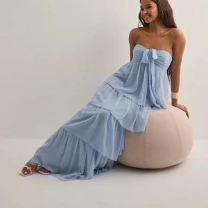 Oanvänd ljusblå volangklänning med rosett - Superfin långklänning i ljusblått från Nelly. Klänningen har flera lager volanger, är axelbandslös och har en söt rosettdetalj framtill. Luftigt material som känns perfekt för varma dagar. Passar dig som gillar romantisk och flowy stil.