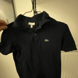 Mörkblå Lacoste pikétröja - Snygg mörkblå pikétröja från Lacoste med klassisk krage och korta ärmar. Ikoniska gröna krokodilloggan på bröstet. Tillverkad i bomull för en skön och luftig känsla. Perfekt för en clean och sportig look. Har använts några gånger, mena har inga som helst slitningar! Köptes för 849kr och jag har växt ur den så vill gärna bli av med den. Passar personer från 160-170 centimeter långa och är rätt slim i passformen!