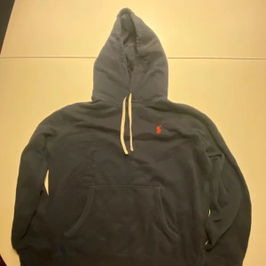 Mörkblå hoodie från Polo Ralph Lauren - Mörkblå hoodie från Polo Ralph Lauren med klassisk röd logga på bröstet. Tröjan har huva med vita snören och en stor magficka. Perfekt för dig som gillar en stilren och avslappnad look. 170/92A