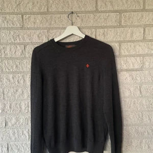 Svart merinoullströja från Morris - Snygg mörkgrå stickad tröja från Morris i 100% merinoull. Klassisk modell med rund halsringning och lång ärm. Diskret röd broderad logga på bröstet ger en stilren touch. Perfekt under kyliga dagar.