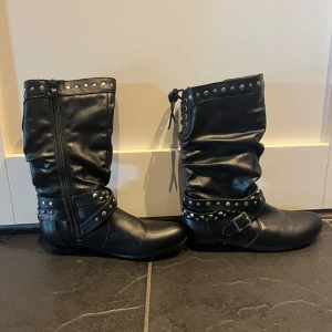 Svarta boots med nitar och leopardfoder - Snygga svarta boots i syntet med coola nitar och spännen runt ankeln. Dragkedja på sidan och snörning bak. Insidan är fodrad med mjuk, rosa leopardmönstrad päls 