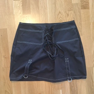 Svart kortkjol med snörning från SHEIN - Svart kortkjol från SHEIN Curve med cool snörning framtill och vita kontrastsömmar. Kjolen har två dekorativa band med metallringar längst ner och är gjord i ett slätt syntetmaterial. Perfekt för dig som gillar edgy detaljer och streetstyle.