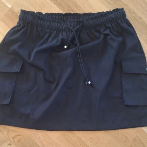 Svart kortkjol från SHEIN Curve - Svart kortkjol från SHEIN Curve med elastisk midja och snörning. Kjolen har två stora fickor på sidorna som ger en cool utility-vibe. Tillverkad i 100% polyester och passar perfekt för dig som gillar en avslappnad men trendig stil.