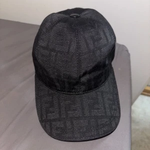Svart Fendi keps - Snygg svart keps från Fendi med diskret FF-monogram över hela ytan. Kepsen har klassisk form, böjd skärm och justerbar rem bak. Tillverkad i ett glansigt tyg som ger en lyxig känsla. Perfekt för dig som vill ha en stilren accessoar med designer-vibe.