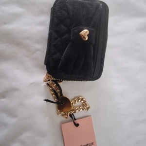 Svart plånbok från Juicy Couture - Liten svart plånbok från Juicy Couture med quiltad framsida i sammet, rosett och guldfärgat hjärtdetalj. Baksidan har kortfack och hjärtformat fönster. Kedja i guld och dragkedja runt om. Perfekt för kort och småsaker.