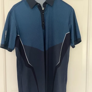  Galvin Green golfpike  - Snygg golfpike från Galvin Green i blå och mörkblå nyanser med vit detalj längs sidorna. Tröjan har klassisk krage, knappar framtill och korta ärmar. Tillverkad i polyester för en luftig känsla. Hör av dig vid funderingar 👍