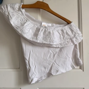 Vit offshoulder topp från Gina Tricot - Superfin vit offshoulder topp från Gina Tricot med bred volangkant i broderad spets och ribbad bomull. Toppen är ärmlös och har en romantisk vibe med blommiga detaljer på volangen. Perfekt för sommarens alla tillfällen!