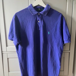 Lila pikétröja från Polo Ralph Lauren - Snygg lila pikétröja från Polo Ralph Lauren med klassisk krage och två knappar framtill. Tröjan har korta ärmar och den ikoniska broderade loggan i turkos på bröstet. Tillverkad i bomull för en skön och luftig känsla.