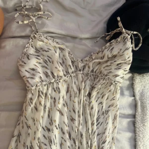 Vit mönstrad sommarklänning från Marina - Supersöt vit klänning med abstrakt mönster i grått och guld från en butik i spanien, Marina. Klänningen har smala axelband med knyt, volangdetaljer och en luftig passform. Perfekt för varma dagar och ger en fräsch och trendig look.