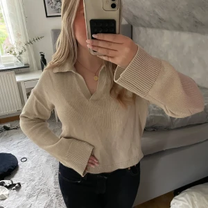 Beige stickad tröja från ONLY - Beige stickad tröja från ONLY med krage och v-ringning. Tröjan har breda ribbade muddar vid ärmslut och nederkant, samt en lite boxig och avslappnad passform. Perfekt för lager-på-lager under hösten och vintern.