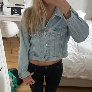 Ljusblå croppad jeansjacka - Säljer en ljusblå croppad jeansjacka med klassisk krage och silverfärgade knappar framtill. Jackan har två bröstfickor med lock och långa ärmar. Perfekt för dig som gillar en chill och trendig stil.