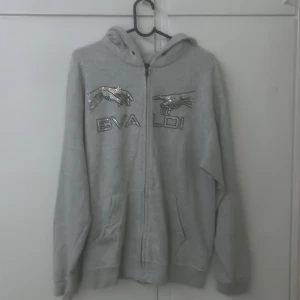 Rhinestone Hoodie - Grå hoodie från BVALDI med glittrande strassdetaljer på bröstet i form av händer och text. Tröjan har huva, dragkedja framtill och två fickor. inte så jätte använd passar s också men lite mer oversize så