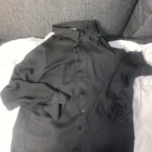 Svart sidenskjorta - Stilren svart skjorta med klassisk krage och knäppning framtill. Skjortan har långa ärmar med manschett och en mjuk, glansig yta som ger en lyxig känsla. Perfekt för dig som gillar en clean och tidlös stil.