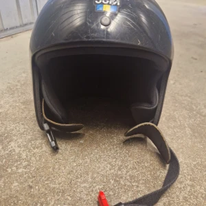 Svart Jofa hjälm med hakrem - Jag skulle uppskatta hjämen från åldern 5-9 år Klassisk svart hjälm från Jofa med rundad form och justerbar hakrem. Utsidan är i hårdplast och insidan är vadderad för extra komfort. Hjälmen har Jofa-logga och svensk flagga på sidan. 