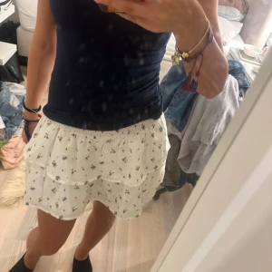 Vit blommig volangkjol med resår - Supersöt vit kortkjol med små blå och gula blommor. Kjolen har volanger och resår i midjan för en bekväm passform. Perfekt för varma dagar och ger en fräsch och somrig look.